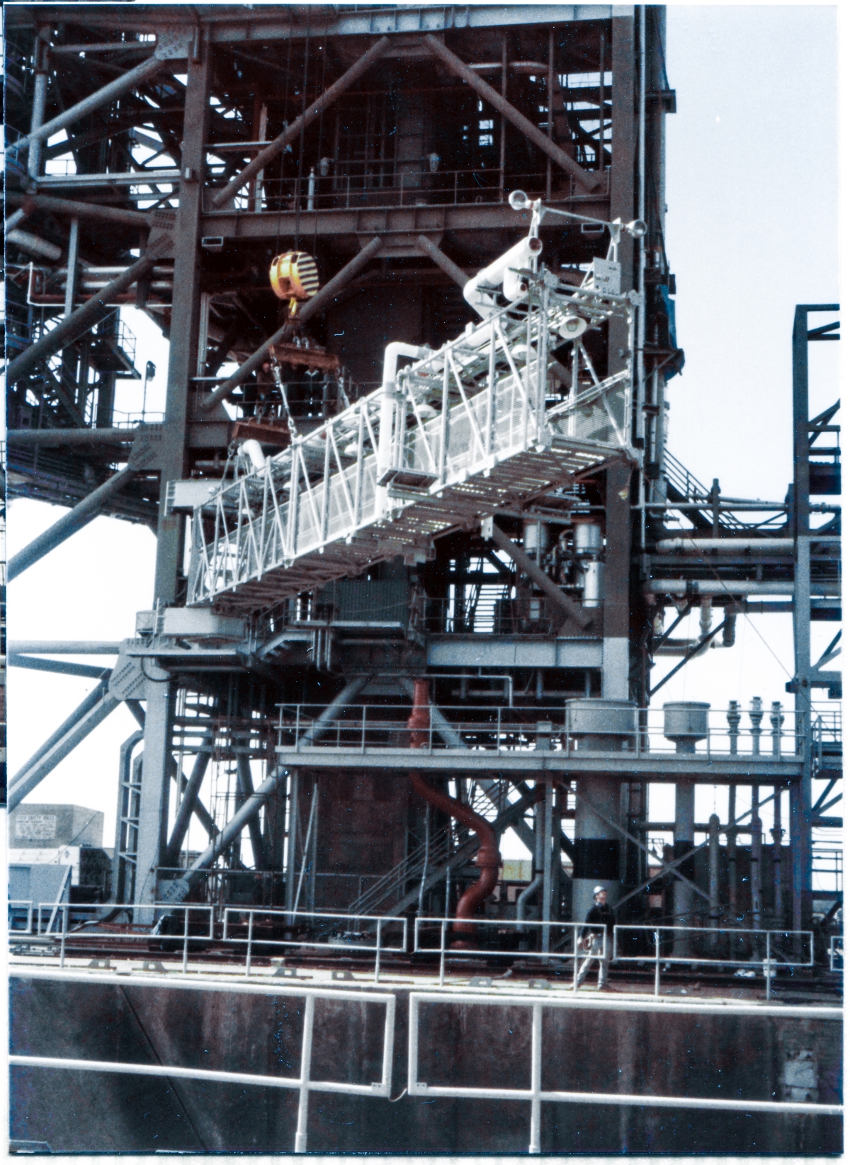 Image 102. At Space Shuttle Launch Complex 39-B, Kennedy Space Center, Florida, the GOX Arm has been raised far enough above the adjacent MLP Utilities Interface Platform by the Hammerhead Crane, to permit Union Ironworkers working for Ivey Steel to begin rotating it around to an orientation more closely matching that which it will assume when it gets bolted to the Strongback near the top of the Fixed Service Structure, at which point the Lift will proceed. The hook on the Hammerhead Crane swivels to permit this reorientation, while the load block which the hook hangs from the bottom of remains in essentially the same position for the duration of the Lift. Photo by James MacLaren.